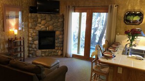 A cozy living room with a stone fireplace, a flat-screen TV, and a dining area with a sink and chairs. - Riverfront condo #3 w/patio, 1block 2 town,King bed,Wifi,20 min ski Granby Ranch (GRAND LAKE)