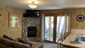 A living room with a stone fireplace, a television mounted above it, and a dining area with a table and chairs. - Riverfront condo #3 w/patio, 1block 2 town,King bed,Wifi,20 min ski Granby Ranch (GRAND LAKE)