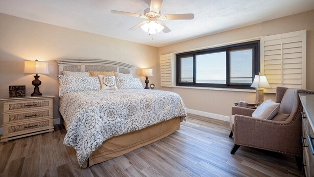 A bedroom with a bed, nightstands, a chair, a ceiling fan, and a window. - Gorgeous 3/3 Oceanfront at Seychelles -- Unit 504 (Daytona Beach Shores)
