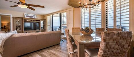 A spacious living area with a large sofa, a dining table with wicker chairs, a ceiling fan, and a chandelier.