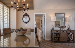 A dining area with a glass table, wicker chairs, a chandelier, a wall clock, a mirror, and a console table with lamps. - Gorgeous 3/3 Oceanfront at Seychelles -- Unit 504 (Daytona Beach Shores)