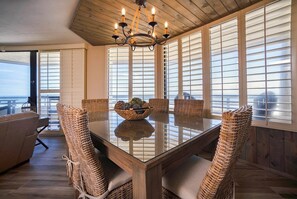 A dining area with a glass table, wicker chairs, a chandelier, and large windows with blinds.