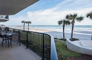 A balcony with a view of the beach, palm trees, and a seating area with chairs and a table.
