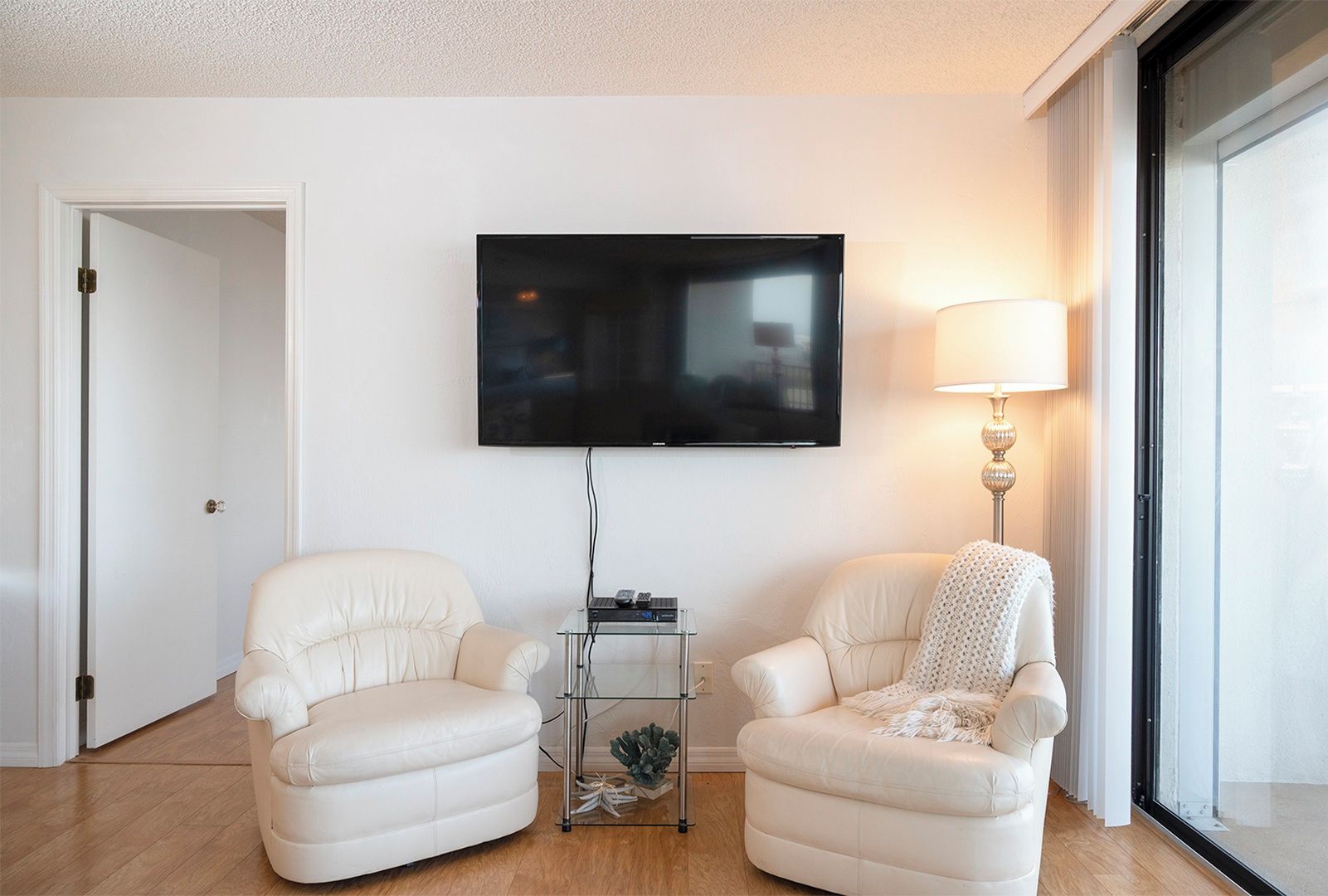 Een woonkamer met twee witte fauteuils, een flatscreen televisie aan de muur, een klein bijzettafeltje met een lamp, en een deur naar een andere kamer.