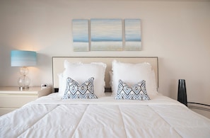 A neatly made bed with white bedding and patterned pillows, a bedside table with a lamp, and a wall with three framed artworks.