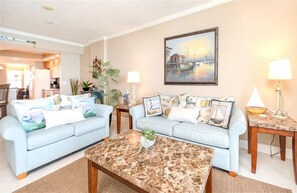 A living room with two light blue sofas, a marble coffee table, and a painting of a harbor scene on the wall.