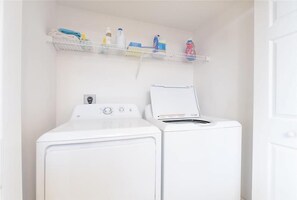 A laundry area with a front-loading washer and dryer, a shelf with cleaning supplies, and a door to the right. - Beautiful Oceanfront 2/2 Condo at Dimucci Twin Towers Unit 1207 (Daytona Beach Shores)