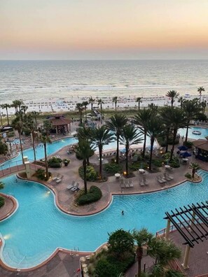 A resort pool area with multiple swimming pools, palm trees, and a beach in the background.