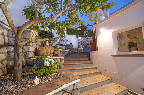 Una escalera de piedra que conduce a un edificio blanco con una ventana, rodeado de un jardín con un árbol y flores.