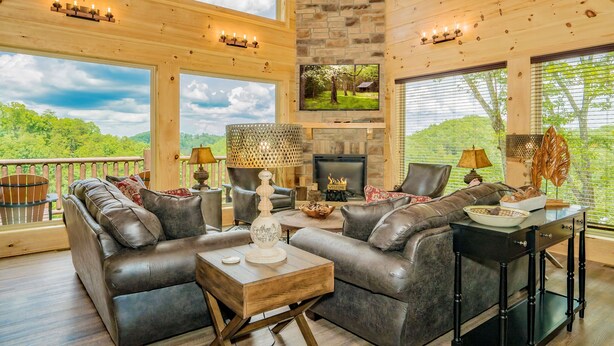 A spacious living room with leather sofas, a fireplace, and large windows overlooking a scenic landscape. - 11 Miles to Gatlinburg! Indoor Heated Pool/Theater Room/Hot Tub/ DISCOUNTS! (Sevierville)