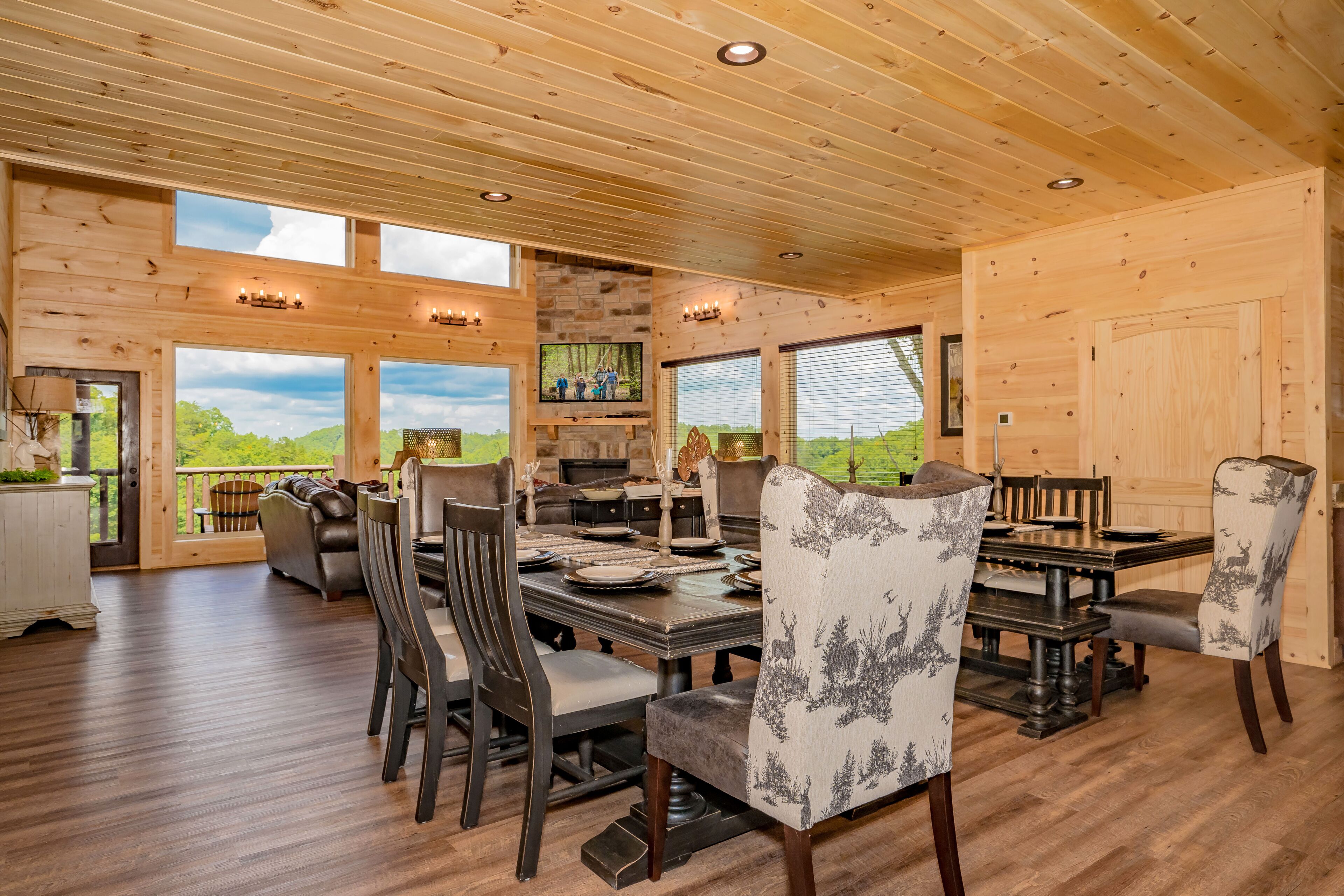 A wooden cabin interior with a dining area, large windows, and a stone fireplace.