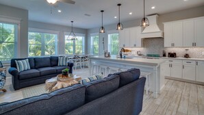 A modern kitchen with white cabinets, a central island, pendant lighting, and a dining area with a sofa and chairs. - EV Charger, Portside Paradise by SeaBreeze Vacation (Holmes Beach)