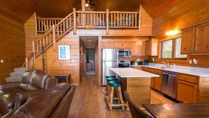 A wooden cabin interior with a living area, kitchen, and a staircase leading to a loft.