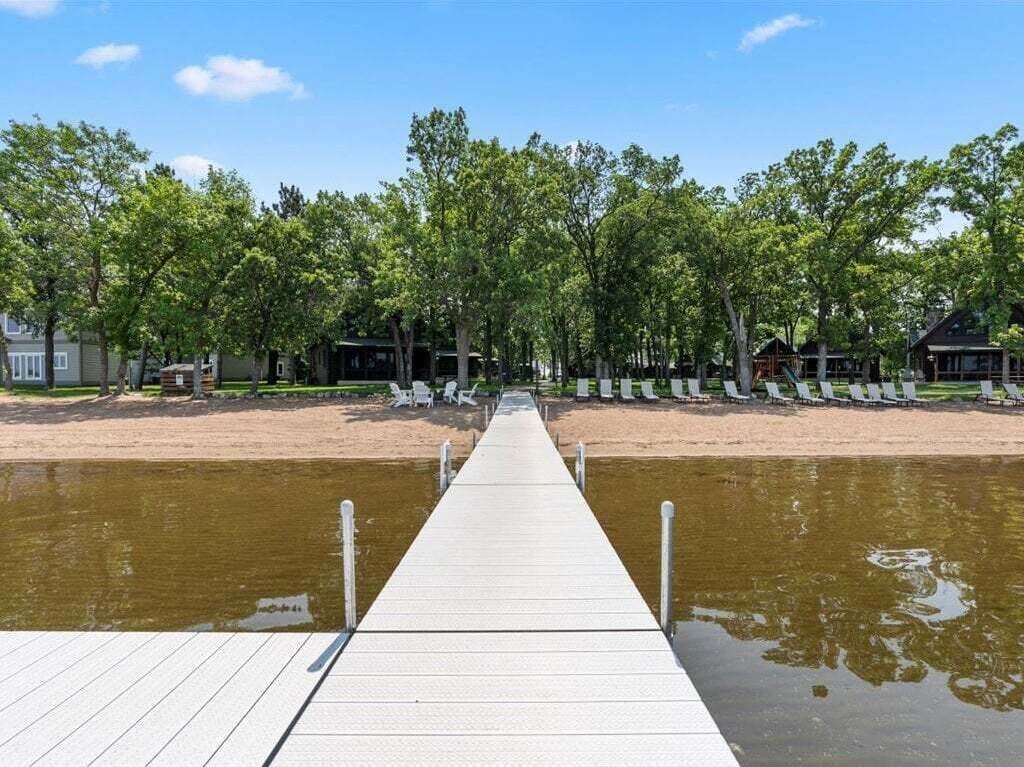 A wooden pier leading to a sandy beach with trees and beach chairs.