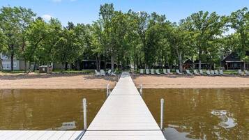 A wooden pier leading to a sandy beach with trees and beach chairs.