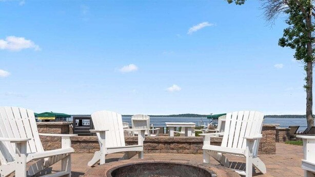 A fire pit surrounded by white Adirondack chairs, with a view of the water and hills in the background. - Lakeside Comfort in the Heart of Gull Lake, Beach Access, Boat Rentals (Nisswa)