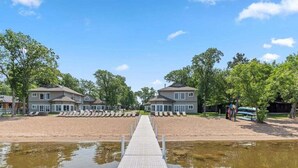 A waterfront property with a dock leading to a sandy beach area, surrounded by trees and two large houses. - Lakeside Comfort in the Heart of Gull Lake, Beach Access, Boat Rentals (Nisswa)
