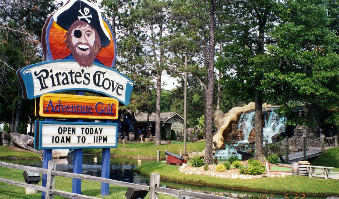 A sign for Pirate's Cove Adventure Golf with a waterfall and wooden structures in the background.