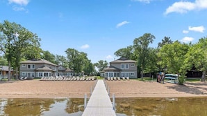 Une propriété située au bord de l’eau, dotée d’un quai menant à une plage de sable, et entourée d’arbres ainsi que de deux grandes maisons.