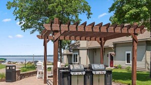 Un pabellón con estructura de pérgola, dos parrillas de barbacoa, mobiliario exterior y vista a un lago.