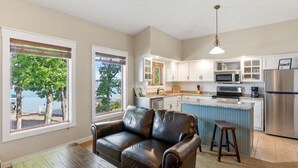 A living room with a leather sofa, wooden coffee table, and large windows with a view of trees and a body of water. - Front Row Seat to Gull Lake Beach | Waterfront Condo (Nisswa)