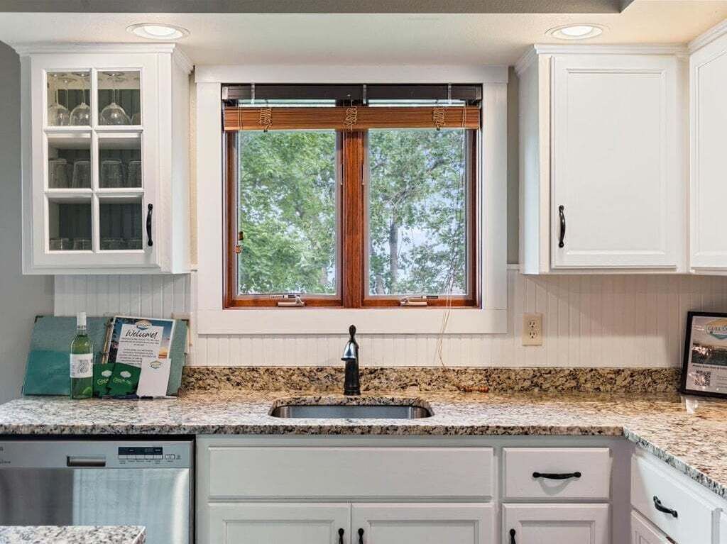 A kitchen with white cabinets, a granite countertop, and a window with a wooden frame.