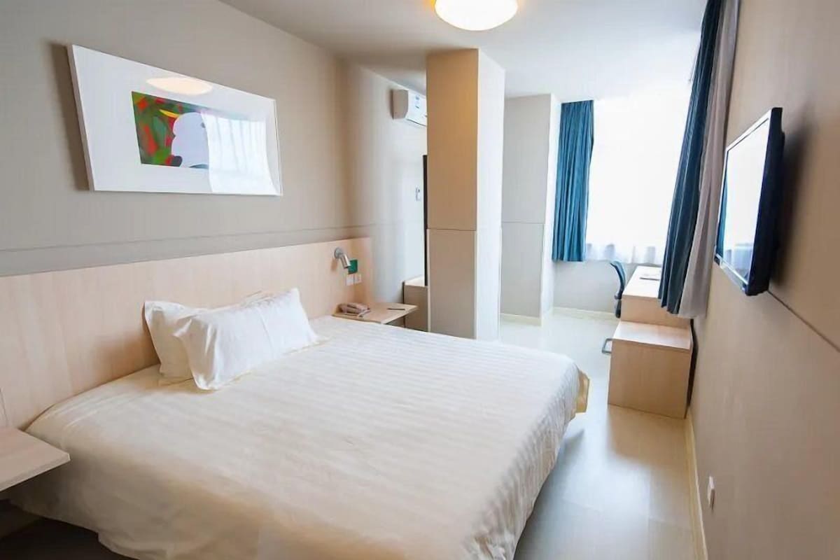Habitació d'hotel amb un llit gran, una taula de nit, un petit escriptori, una cadira, un televisor fixat a la paret i una finestra amb cortines.