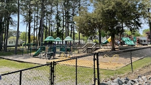 Un parque infantil con toboganes verdes y una zona vallada, rodeado de árboles altos.