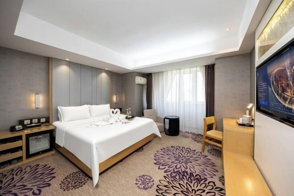 A modern hotel room with a large bed, a desk, a chair, a TV, and a small table with a lamp.