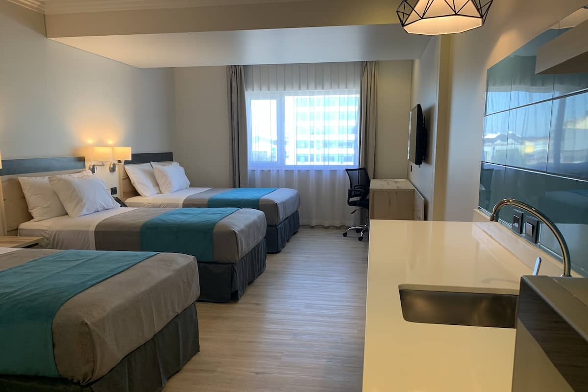 Habitación de hotel con tres camas, un escritorio, una silla, un ventanal grande y un lavamanos moderno.