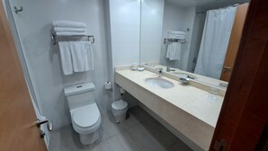 Un baño con inodoro, lavamanos y espejo. Hay toallas en un perchero y un cubo de basura.