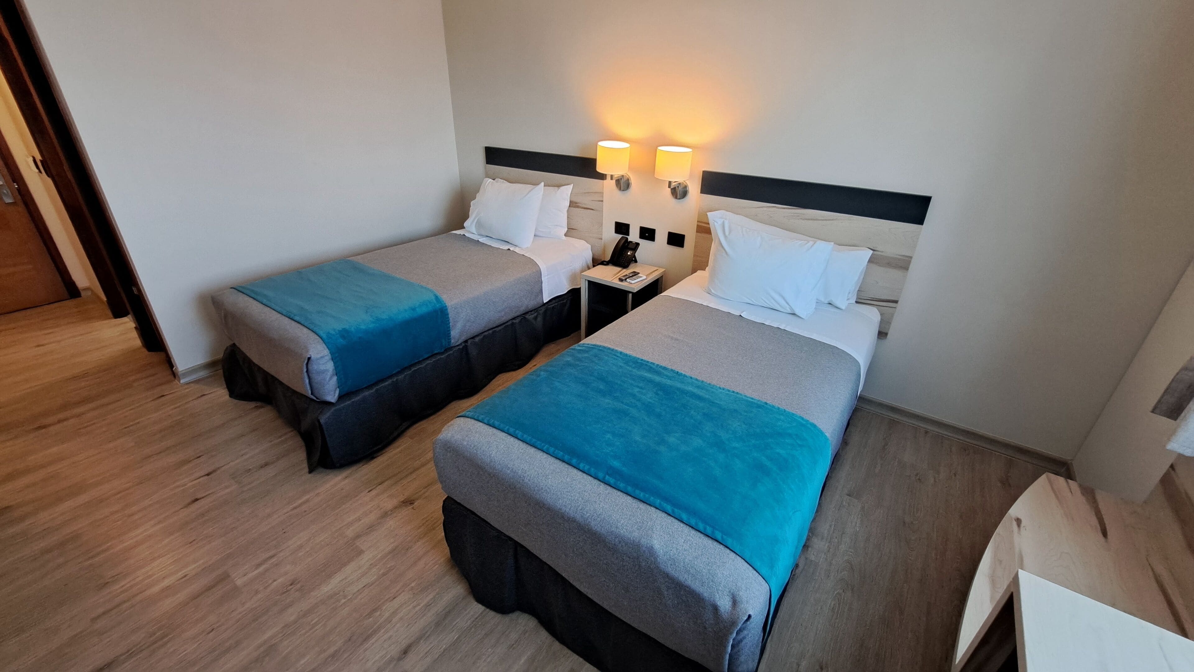 Habitación de hotel con dos camas individuales, cada una con cubrecama azul y gris, almohadas blancas y cabeceros de madera. Se observan mesitas de noche con lámparas y un teléfono.