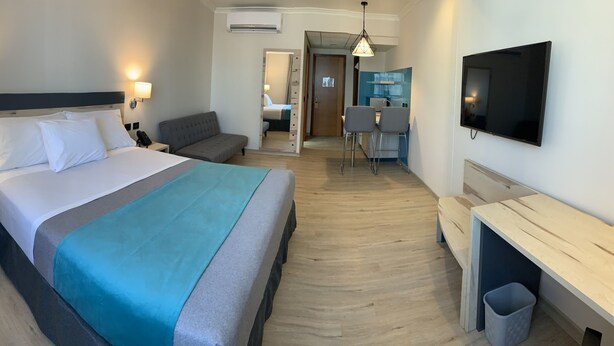 Habitación de hotel con cama, mesitas de noche, escritorio, silla, televisor y kitchenette.