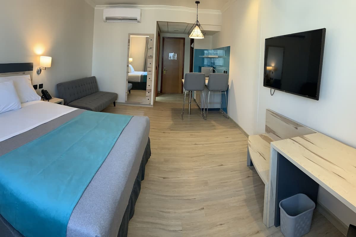 Habitación de hotel con cama, mesitas de noche, escritorio, silla, televisor y kitchenette.