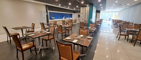 Un comedor amplio con varias mesas y sillas, una zona de cocina al fondo y un ambiente luminoso y bien iluminado.
