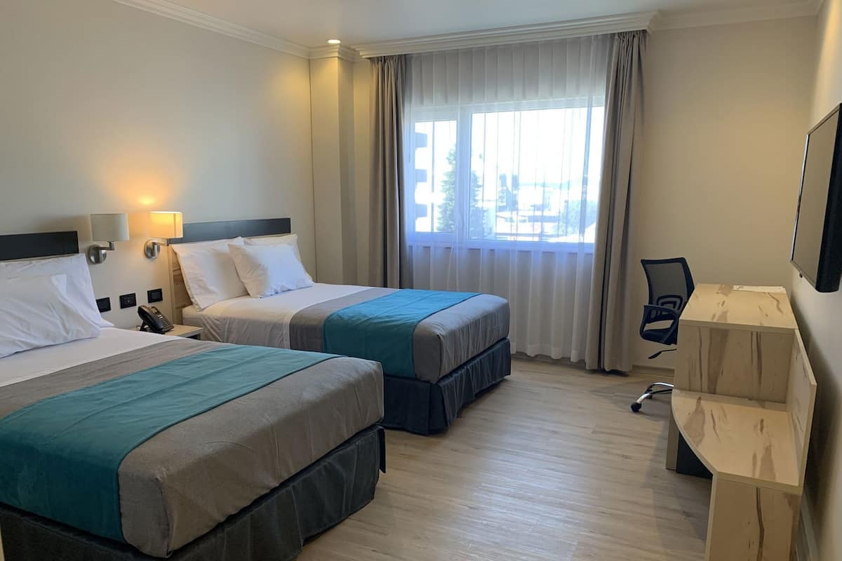 Una habitación de hotel con dos camas, un escritorio, una silla y un ventanal con cortinas.