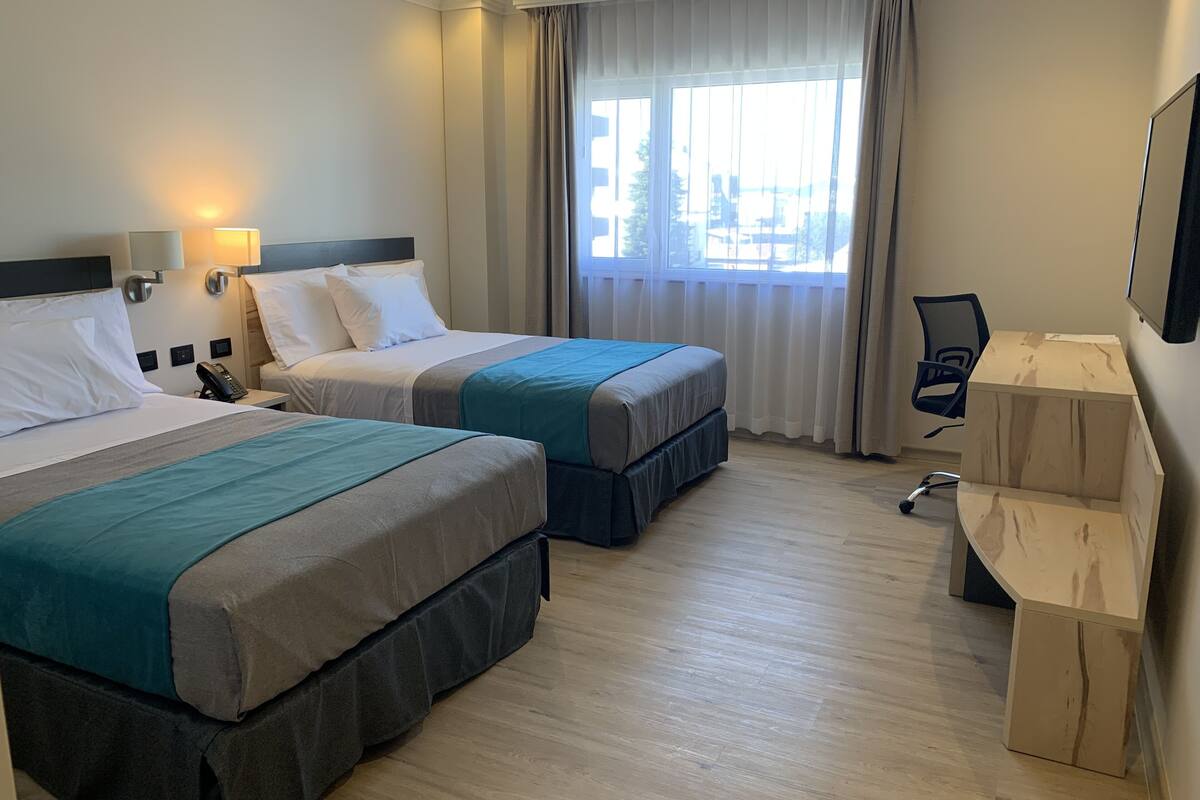 Habitación de hotel con dos camas, un escritorio, una silla y un ventanal con cortinas.