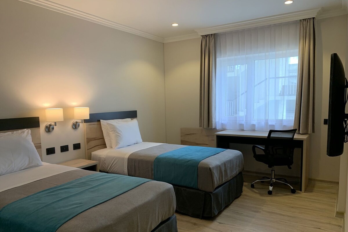 A hotel room with two beds, a desk, a chair, and a window with curtains.
