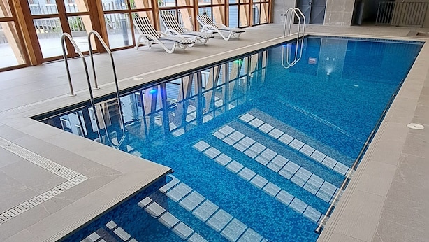 Una piscina interior con agua azul cristalina, vigas de madera y ventanas grandes.