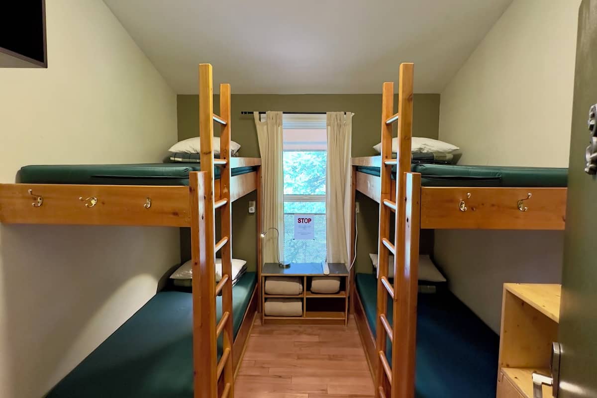 A dormitory room with bunk beds, a small desk, a window with curtains, and a ceiling light.