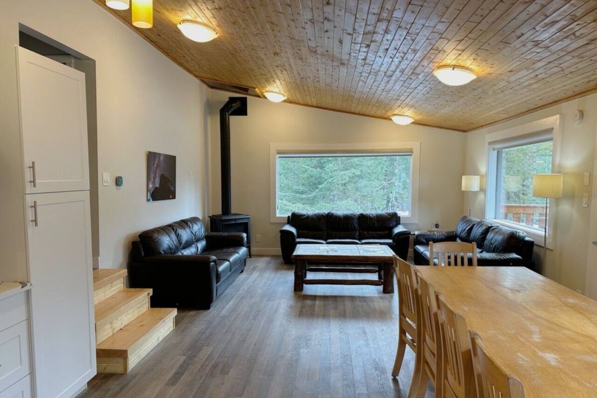 A modern living room with a wooden floor, black leather sofas, a wooden coffee table, and a dining table with chairs. There is a large window providing a view of the outdoors.