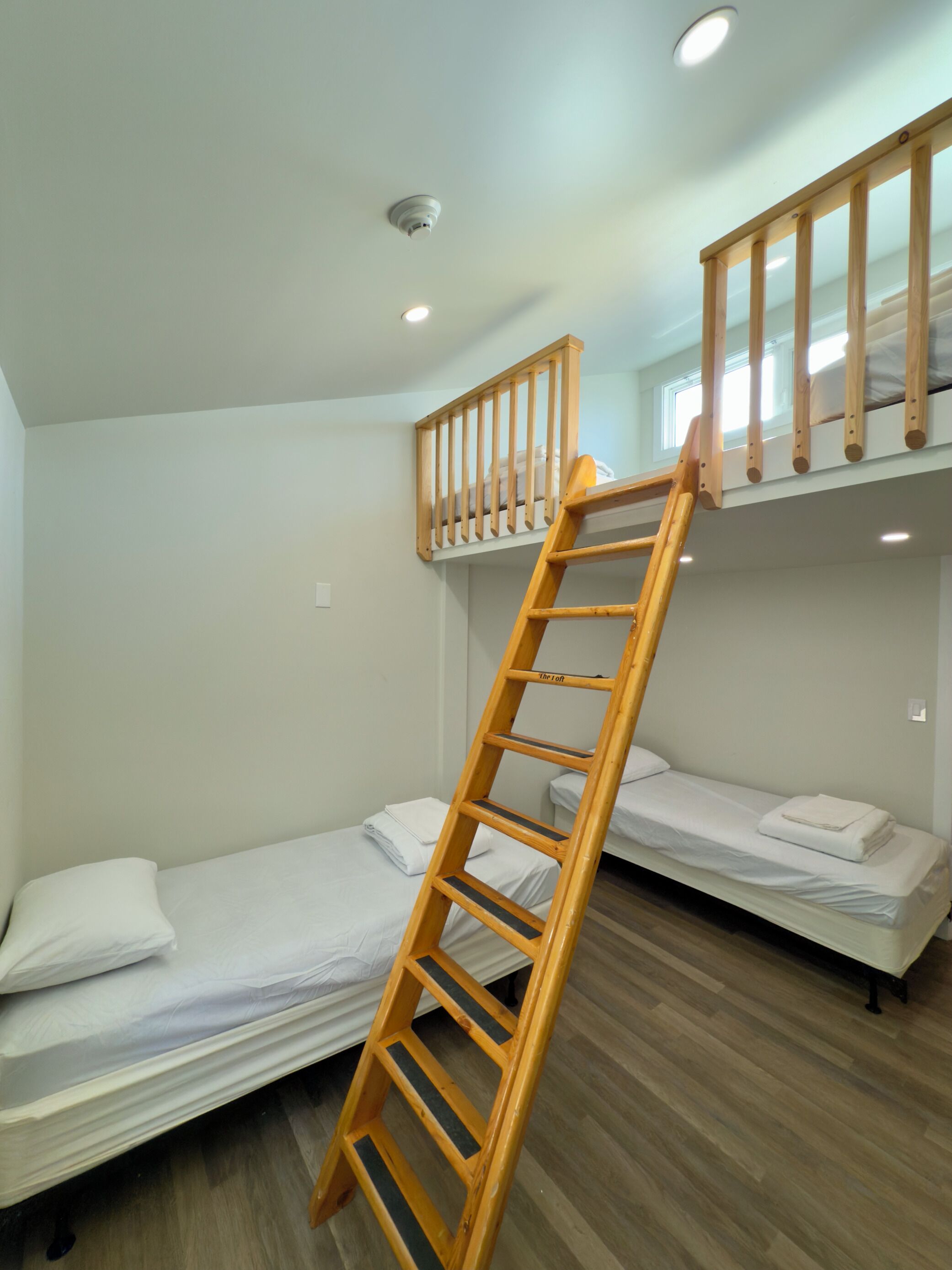 A dormitory room with bunk beds, a wooden ladder, and a window.