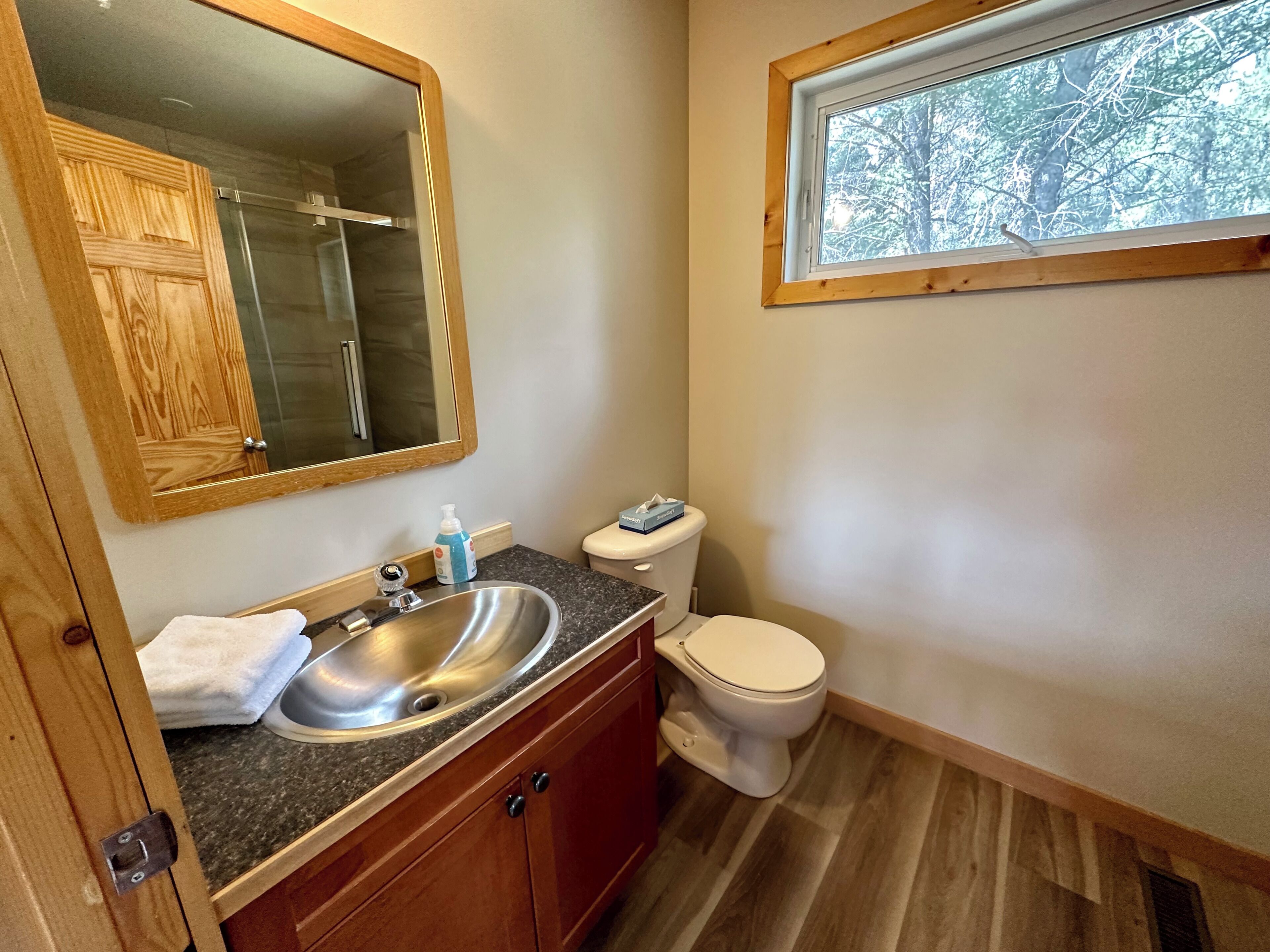 Une salle de bain avec un meuble-lavabo en bois, un évier en acier inoxydable, des toilettes et une fenêtre donnant sur les arbres.