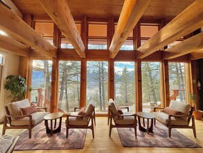 Ein geräumiges Zimmer mit großen Fenstern, Holzbalken und Blick auf einen Wald und Berge.