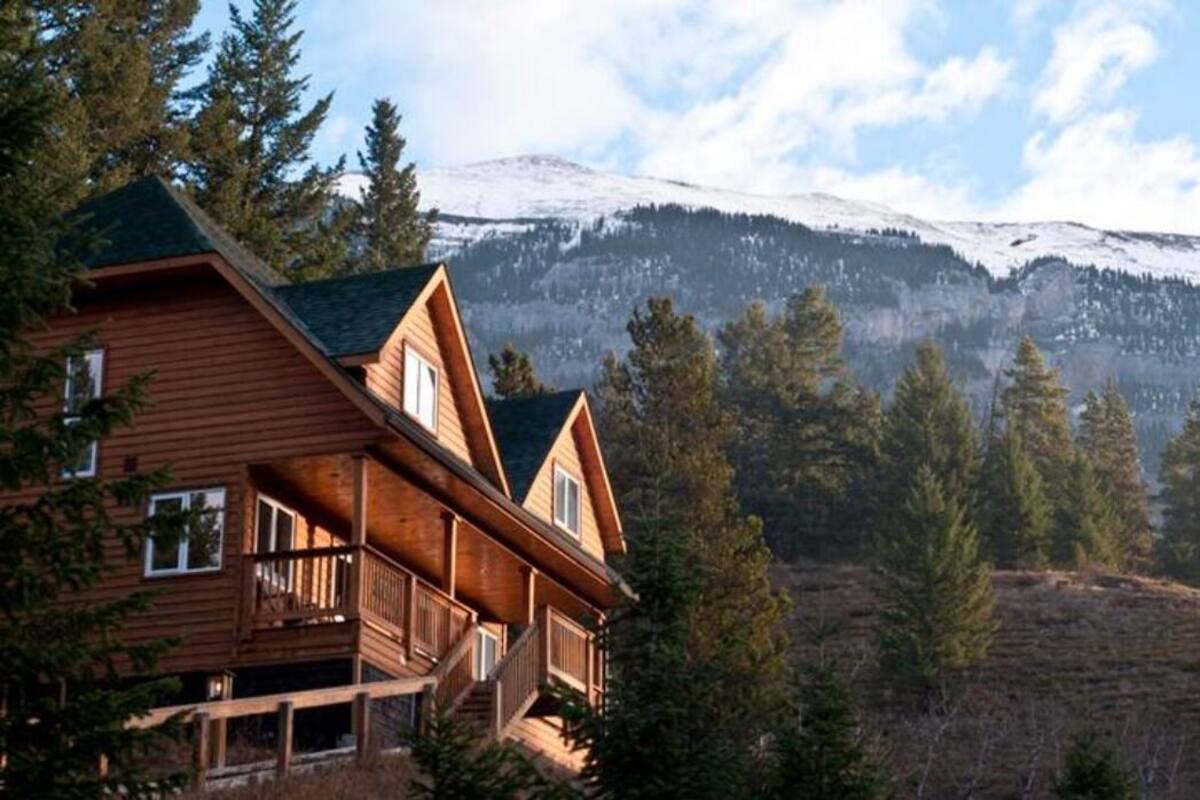 A wooden cabin with a balcony, surrounded by trees and a snow-capped mountain in the background.