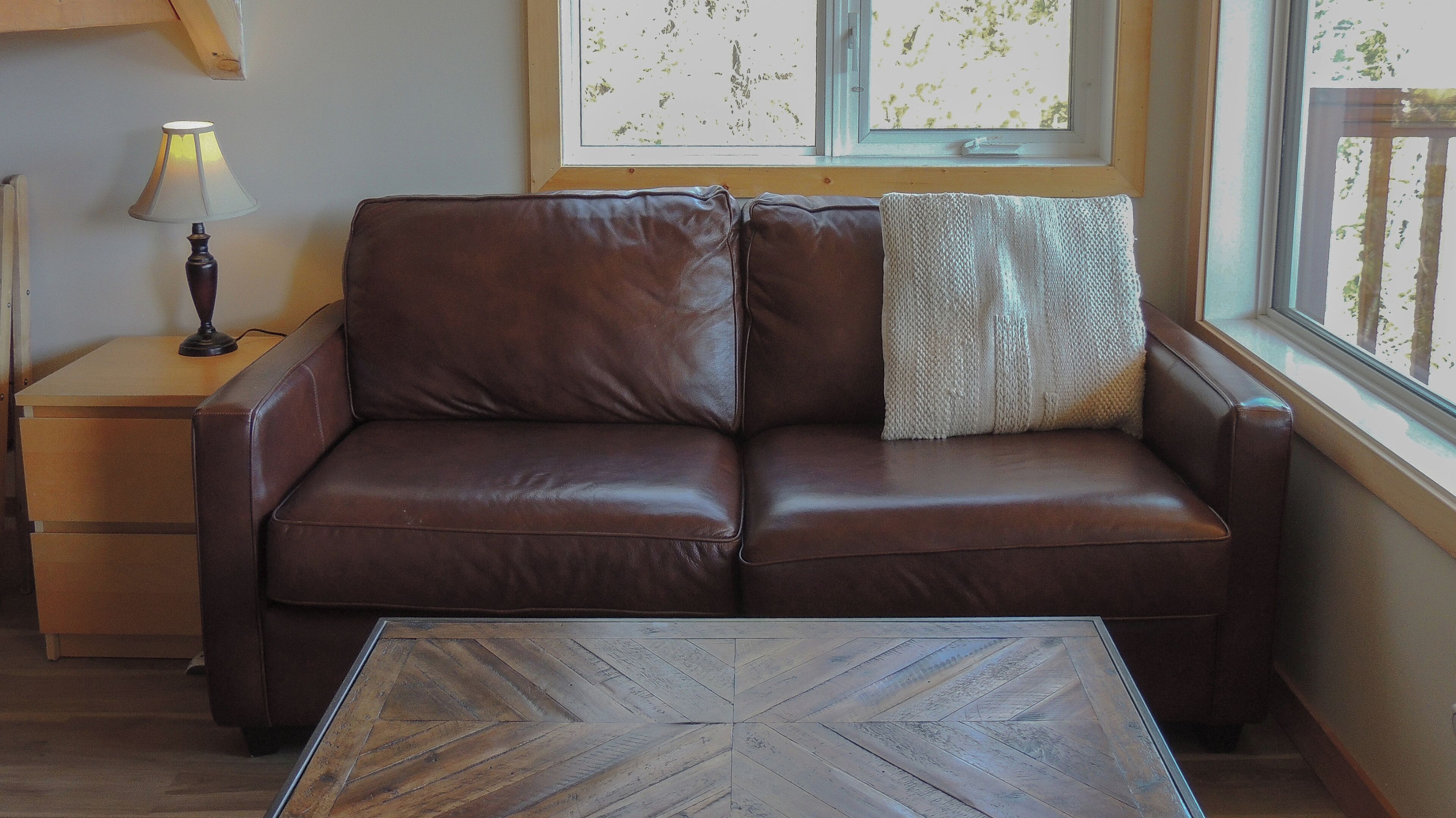 A brown leather sofa with a knitted throw pillow, a wooden side table with a lamp, and a herringbone-patterned coffee table.