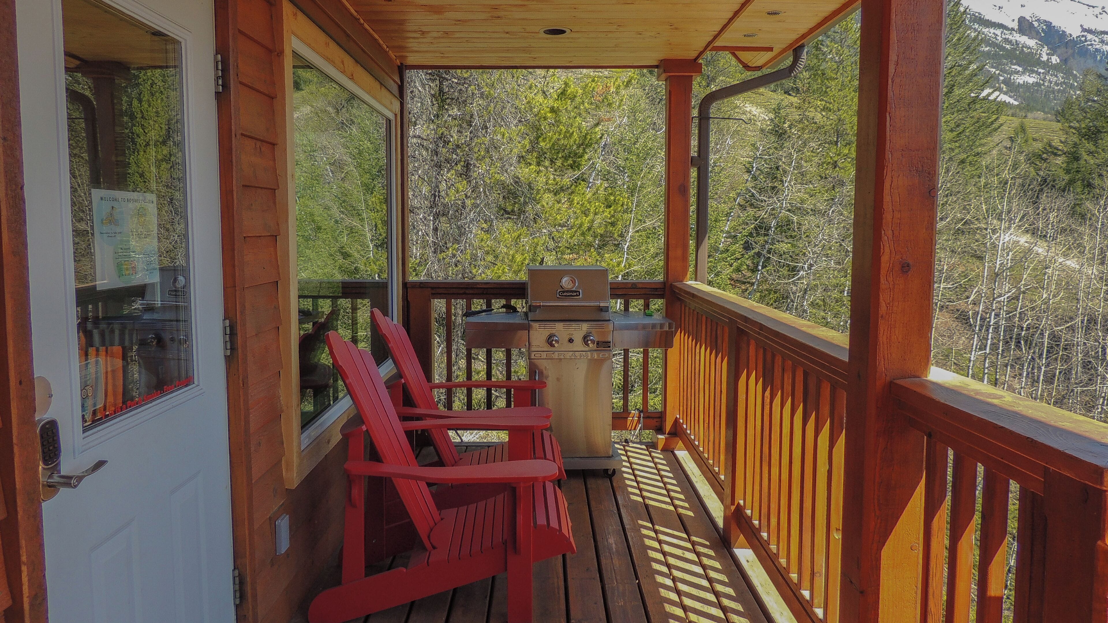 Une terrasse en bois avec un barbecue, des chaises Adirondack rouges et une vue sur une chaîne de montagnes boisées.
