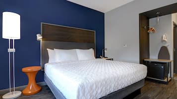 A modern hotel room with a large bed, a bedside table, a lamp, and a small orange stool.