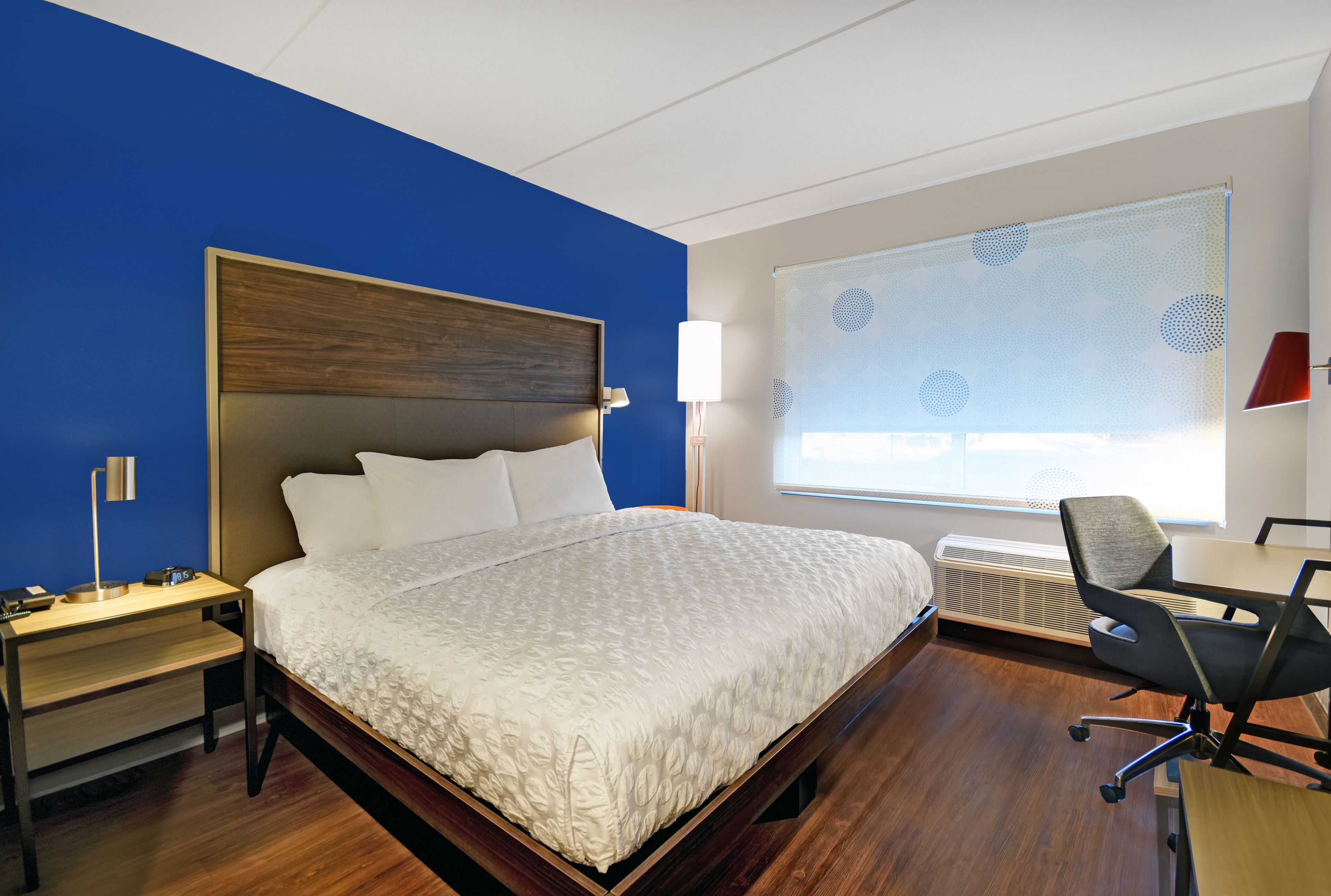 A hotel room with a large bed, a desk with a chair, a nightstand, and a window with blinds.