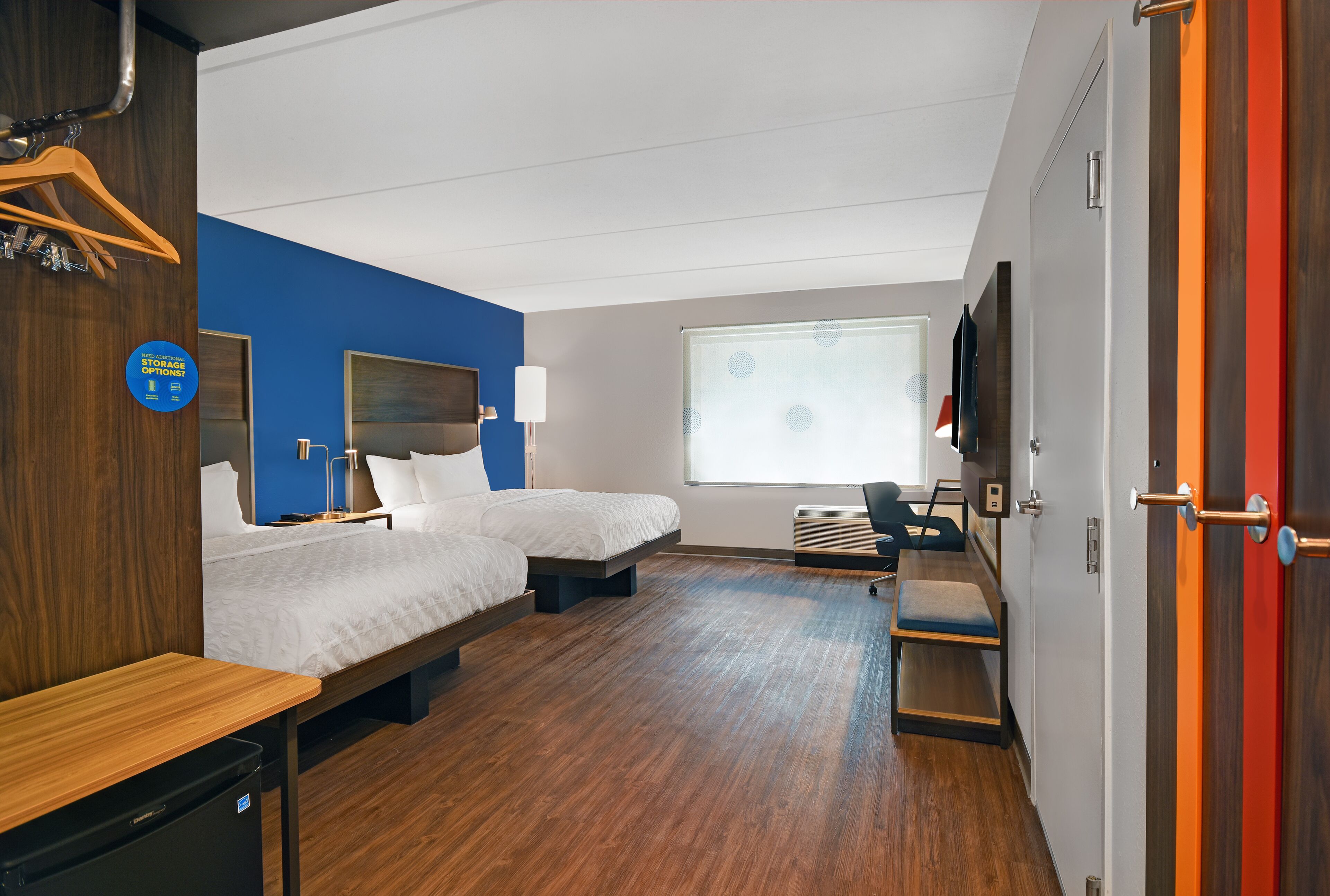 Une chambre d’hôtel avec deux lits, un bureau, une chaise, une télévision et une fenêtre avec des stores.